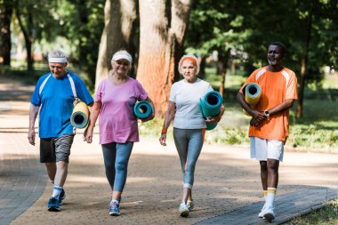 happy senior and multicultural pensioners holding fitness mats and walking in walkway in park  clipart