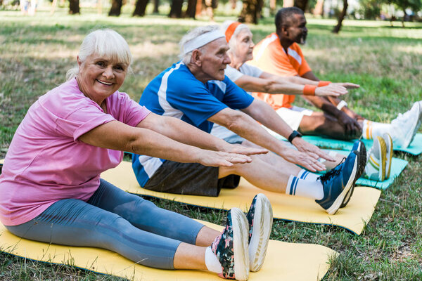 selective focus of happy senior woman in sportswear exercising  near multicultural pensioners on fitness mats 