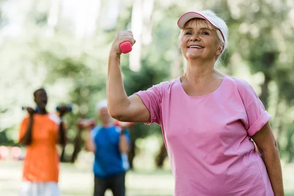 年金受給者の近くで運動しながらダンベルを保持するキャップで幸せな引退した女性の選択的な焦点 — ストック写真