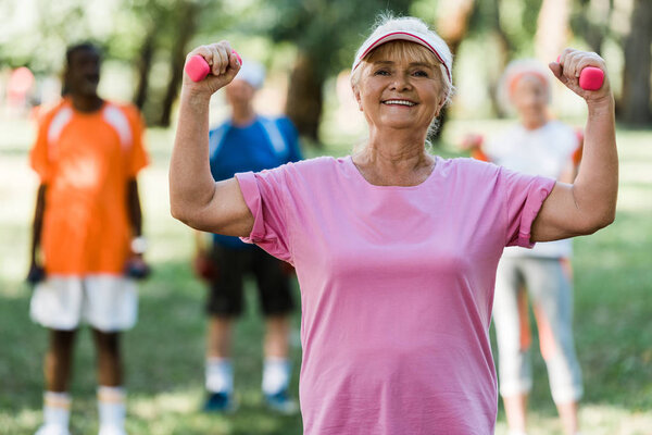 selective focus of happy retired woman in cap holding dumbbells while exercising near pensioners 