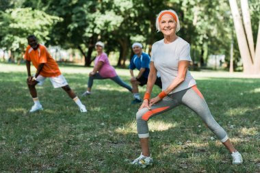selective focus of cheerful retired woman stretching near multicultural pensioners on grass  clipart