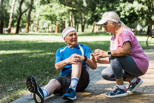 senior man touching knee while sitting on walkway near retired woman in cap 