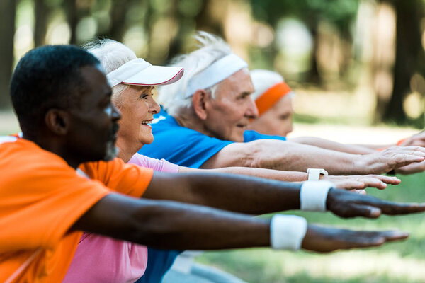 selective focus of happy senior man standing with outstretched hands near multicultural pensioners 