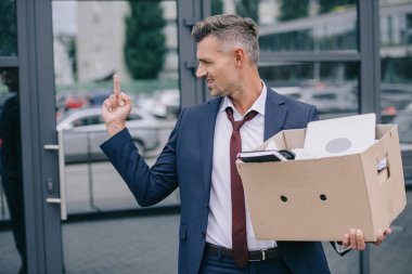 upset man in suit showing middle finger while holding box near building  clipart