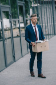 dismissed businessman walking near building and holding carton box 