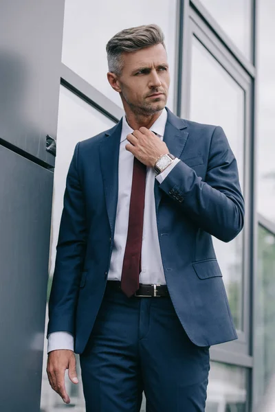 serious businessman in formal wear touching tie outside near building