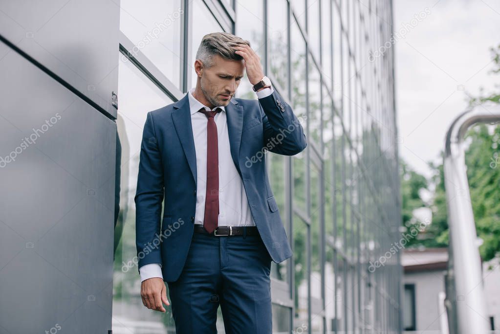 selective focus of upset businessman in formal wear standing near building and touching hair 