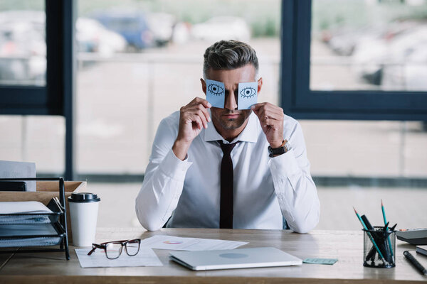 businessman holding sticky notes with drawn eyes near face 