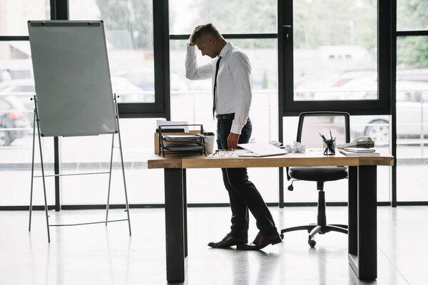 upset businessman touching head while standing near desk in office 