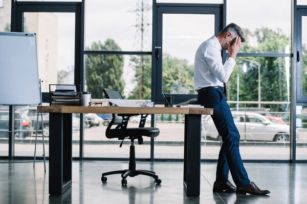 businessman touching head while standing near table in modern office 