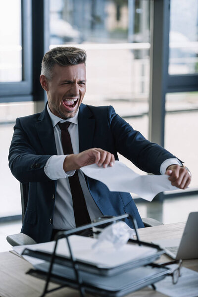 selective focus of irritated man screaming while tearing paper in office 