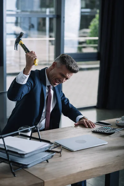 selective focus of irritated businessman holding hammer near laptop on table