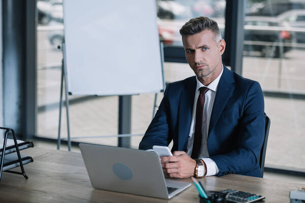 selective focus of handsome man looking at camera while holding smartphone near laptop 