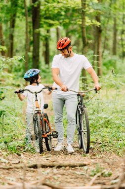 full length view of father and son walking with bicycles in forest clipart