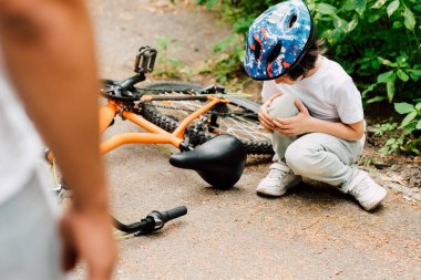 selective focus of boy fell from bicycle and looking at knee while father standing near son clipart