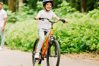 selective focus of happy boy riding bicycle while father standing and looking at son  clipart