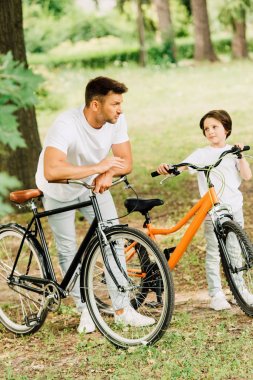 parkta dururken bisiklete yaslanan baba ve oğulun tam boy görünümü