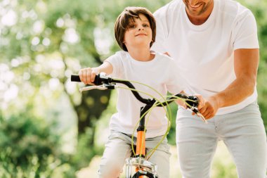 cropped view of father helping son while kid riding bicycle and looking forward  clipart