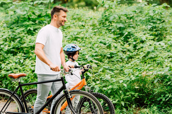 side view of son and father standing with bicycles near forest and looking forward