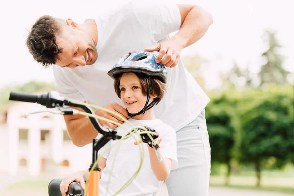 selective focus of father putting helmet on son while boy standing near bicycle and looking away