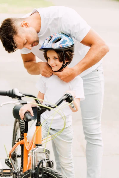 father putting helmet on son while boy standing near bicycle and looking away