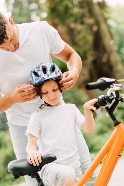 father putting helmet on son while boy trying to sit on bicycle
