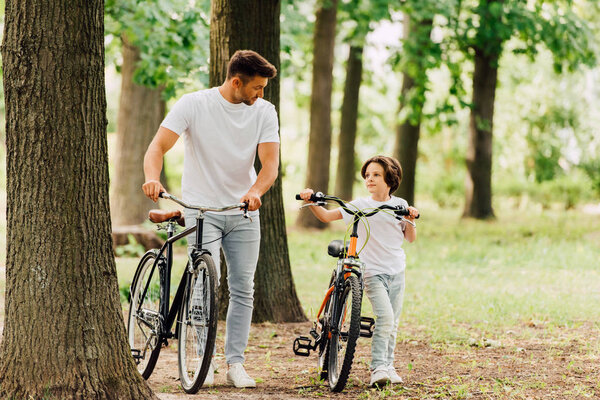 full length view of father and son with bicycles walking in park and looking at each other