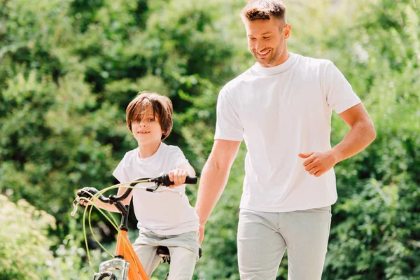 息子は自転車に乗り 父親は子供の隣を歩き 自転車の座りを持っている — ストック写真