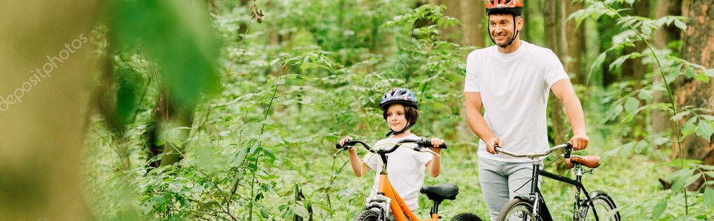 Panoramic shot of father and son in helmets walking with bicycles in forest