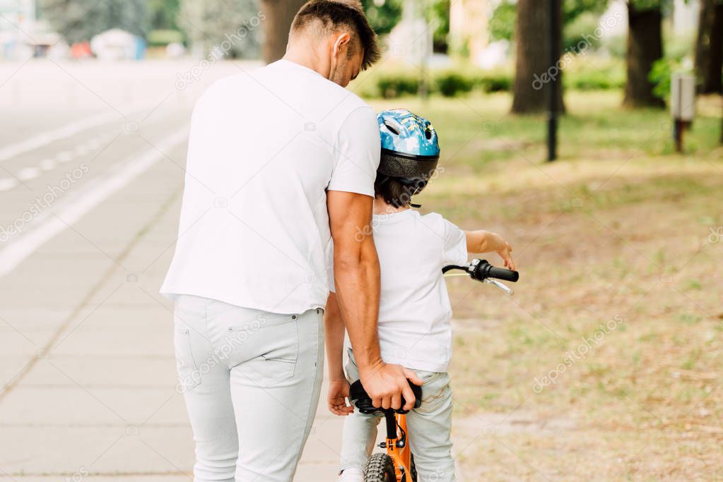 back view of father helping son to ride on bicycle while kid sitting on bike