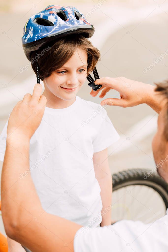 selective focus of boy looking at dad and smiling while father putting helmet on kid