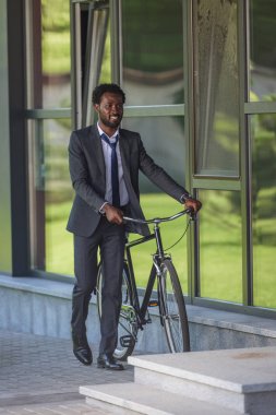 ofis binası yakınında bisikletle yürüyen gülümseyen afrikalı-amerikalı işadamı