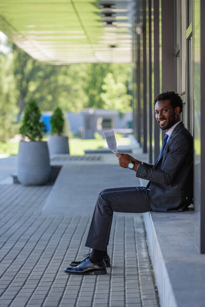 cheerful african american businessman smiling at camera while sitting on parapet and holding newspaper