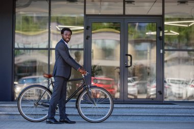 ofis binasıyakınında dururken kameraya gülümseyen neşeli afrikalı-amerikalı işadamı