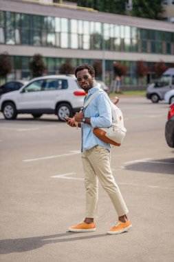 yakışıklı afro-amerikan adam araba park ayakta ve akıllı telefon tutarak kameraya bakıyor