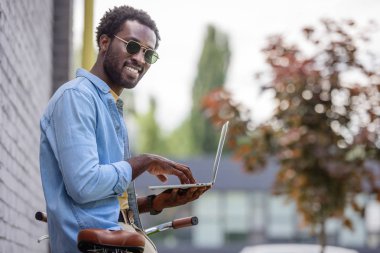 yakışıklı afro-amerikan adam bisiklet yanında duran ve dizüstü bilgisayar kullanarak kameraya gülümseyen