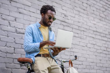 bisiklet yanında tuğla duvar tarafından ayakta iken dizüstü bilgisayar kullanarak yakışıklı afro-amerikan adam