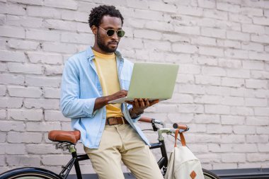 attentive, stylish african american man using laptop while standing by brick wall near bicycle clipart