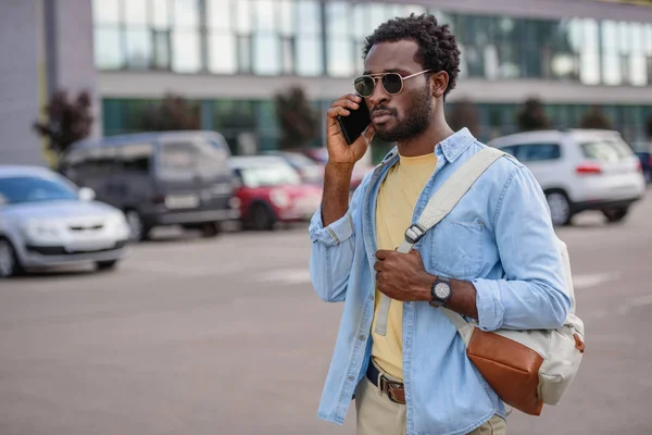 駐車場に立って目をそらしながらスマートフォンで話すハンサムなアフリカ系アメリカ人の男性 — ストック写真