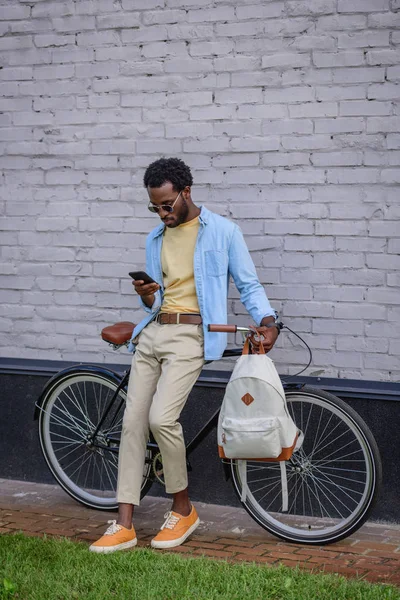 Przystojny African American Man Pomocą Smartfona Stojąc Rowerem Pobliżu Muru — Zdjęcie stockowe