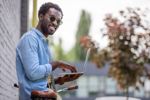 handsome african american man smiling at camera while standing near bicycle and using laptop