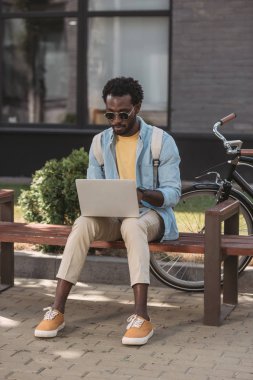 bisiklet yakınındaki bankta otururken dizüstü bilgisayar kullanan şık afro-amerikan adam