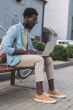 bankta otururken dizüstü bilgisayar kullanarak güneş gözlüğü şık afro-amerikan adam