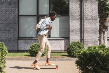 güneşli sokakta longboard sürme sırt çantası ile şık afrikalı-amerikalı adam