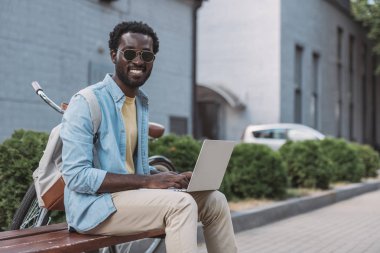 neşeli afro-amerikan adam bankta otururken ve dizüstü bilgisayar kullanarak kameraya gülümseyen