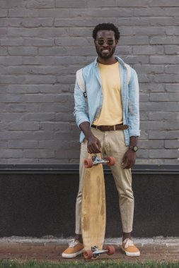yakışıklı afro-amerikan adam longboard ile tuğla duvar yakınında dururken kameraya gülümseyen