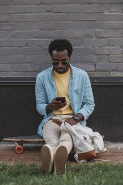 duvara yakın longboard otururken akıllı telefon kullanarak şık afro-amerikan adam