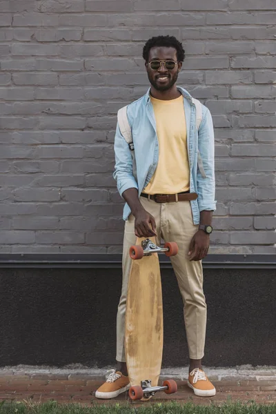 ハンサムなアフリカ系アメリカ人男性が長板でレンガの壁の近くに立っている間 カメラで微笑む — ストック写真