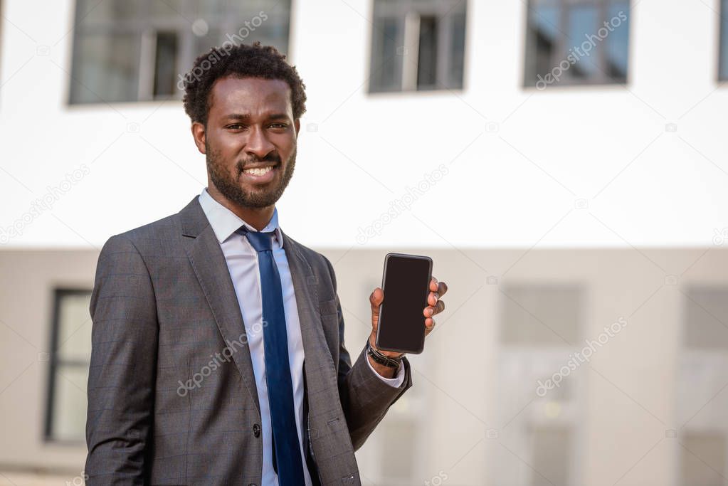 cheerful african american businessman showing smartphone with blank screen and smiling at camera