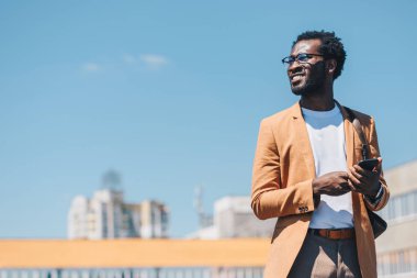akıllı telefon kullanarak neşeli afrikalı amerikalı işadamı ve arka planda mavi gökyüzü ile gülümseyen